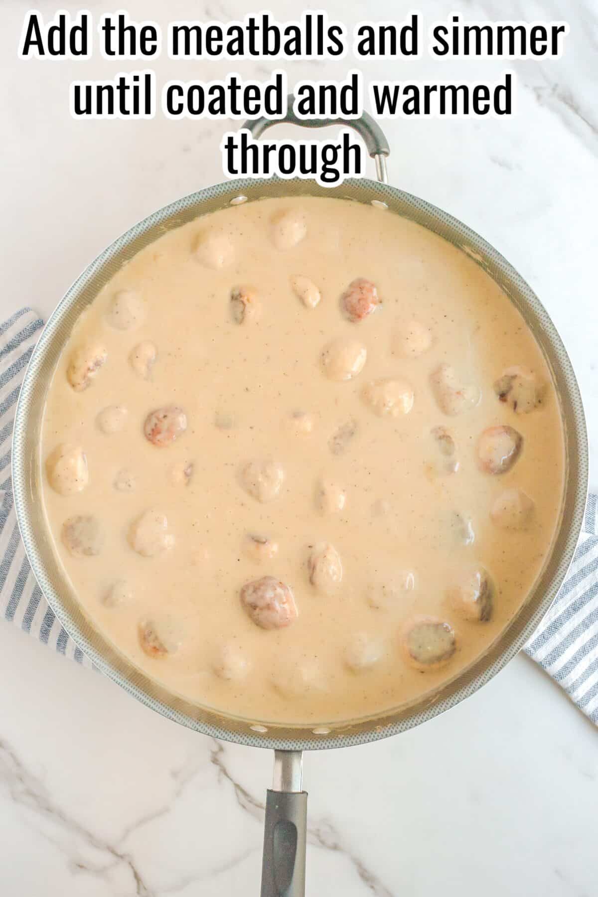 A skillet filled with meatballs simmering in a creamy sauce sits on a marble countertop with a striped cloth underneath.