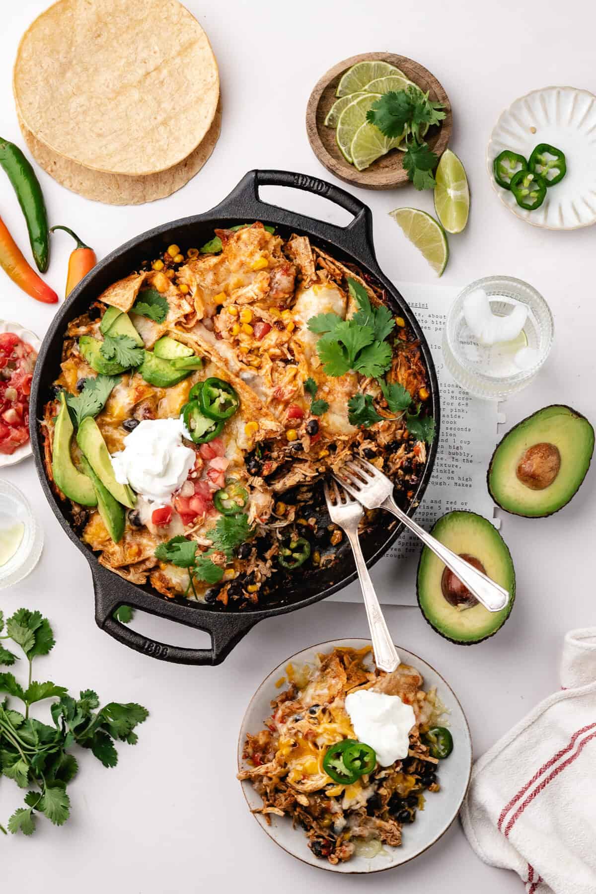 Cast iron skillet filled with chicken enchilada casserole, topped with avocado, jalapeño, cilantro, and sour cream, with a small serving on a side plate.