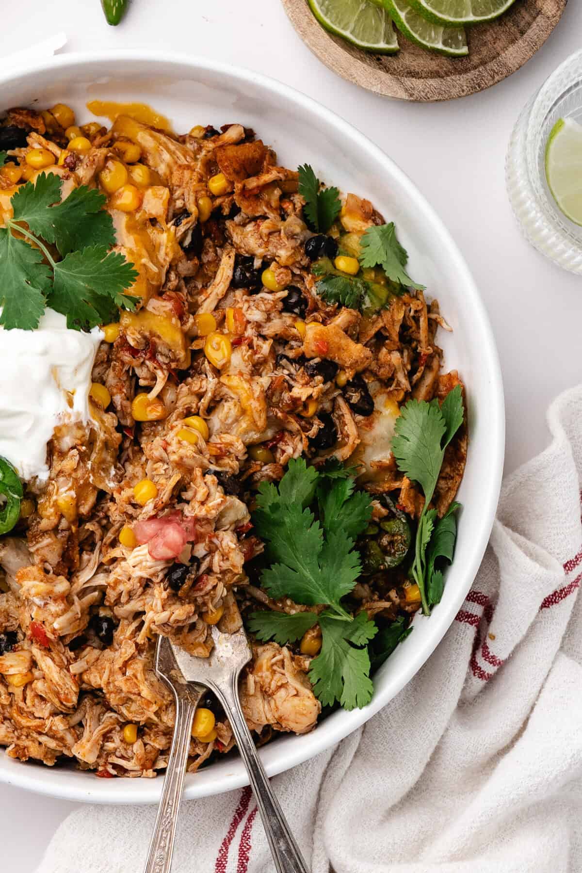 A white bowl of chicken, corn, and black bean casserole topped with sour cream and cilantro, with lime wedges on the side.