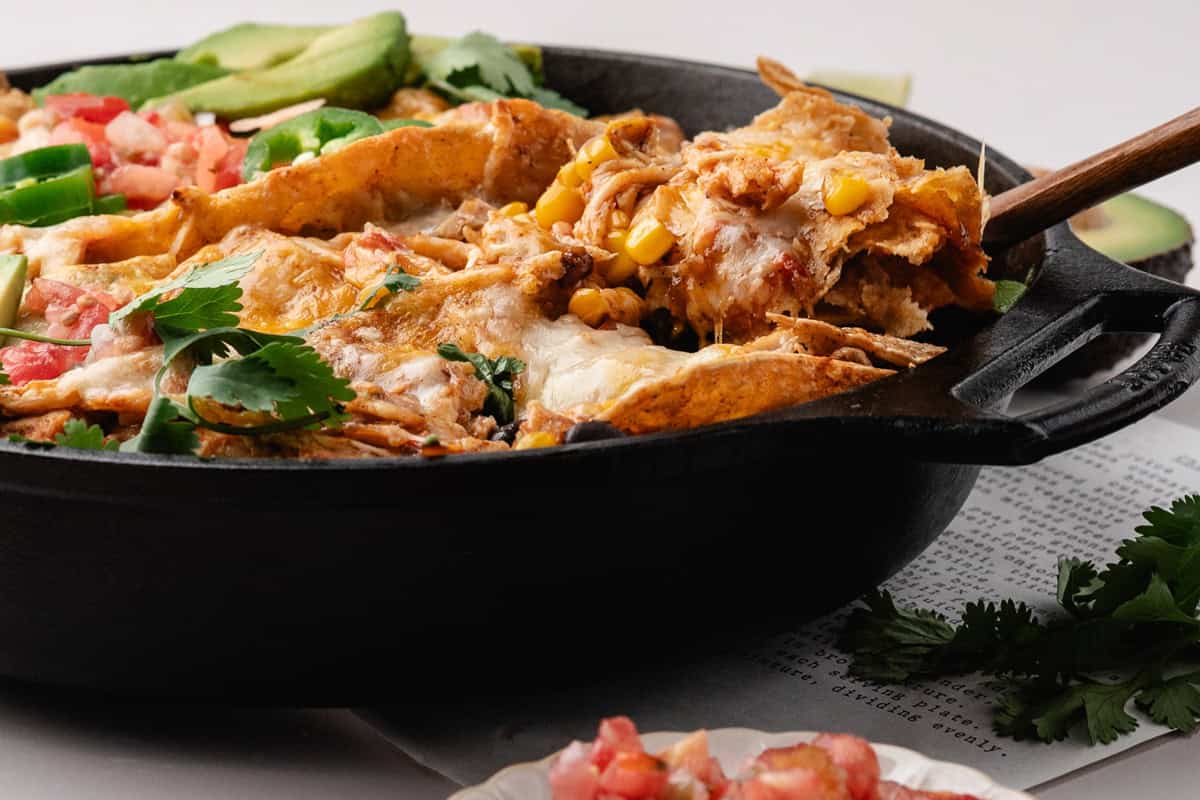 A wooden spoon lifting cheesy chicken enchilada casserole with corn and cilantro from a cast-iron skillet.