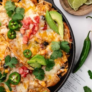 Skillet enchilada casserole topped with melted cheese, diced tomatoes, sliced jalapeños, avocado slices, and cilantro, served with lime wedges and a whole green chili.
