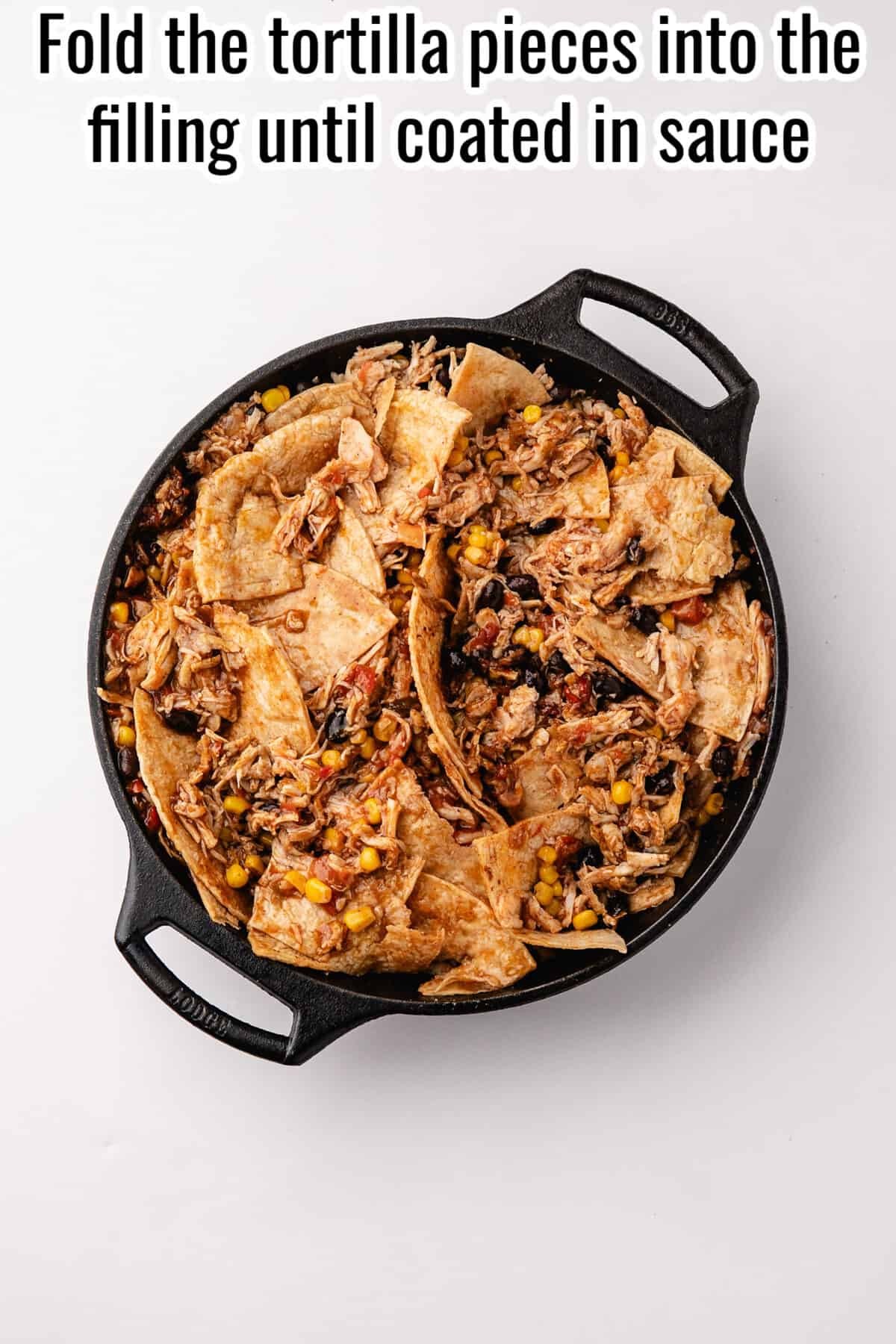 Cast iron skillet containing a mixture of tortilla pieces, shredded chicken, corn, and black beans, with text reading: “Fold the tortilla pieces into the filling until coated in sauce.”