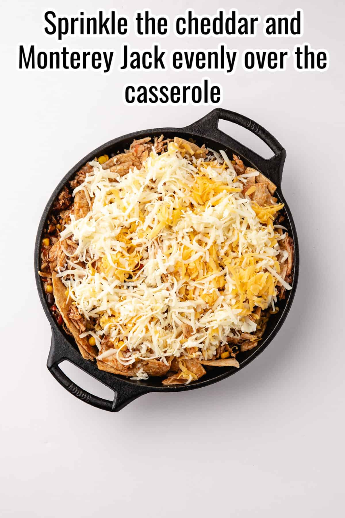A cast iron skillet containing a casserole topped with shredded cheddar and Monterey Jack cheese, with the text “Sprinkle the cheddar and Monterey Jack evenly over the casserole” above it.