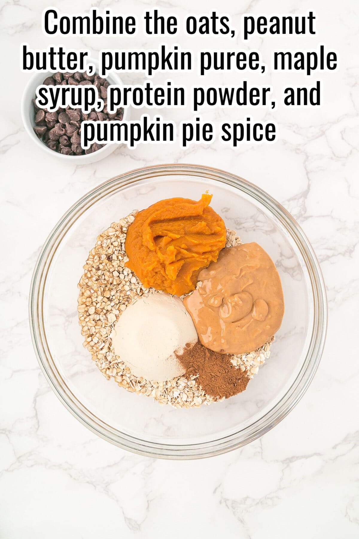 A glass bowl with oats, pumpkin puree, peanut butter, protein powder, and pumpkin pie spice on a marble surface; a small bowl of chocolate chips is nearby.