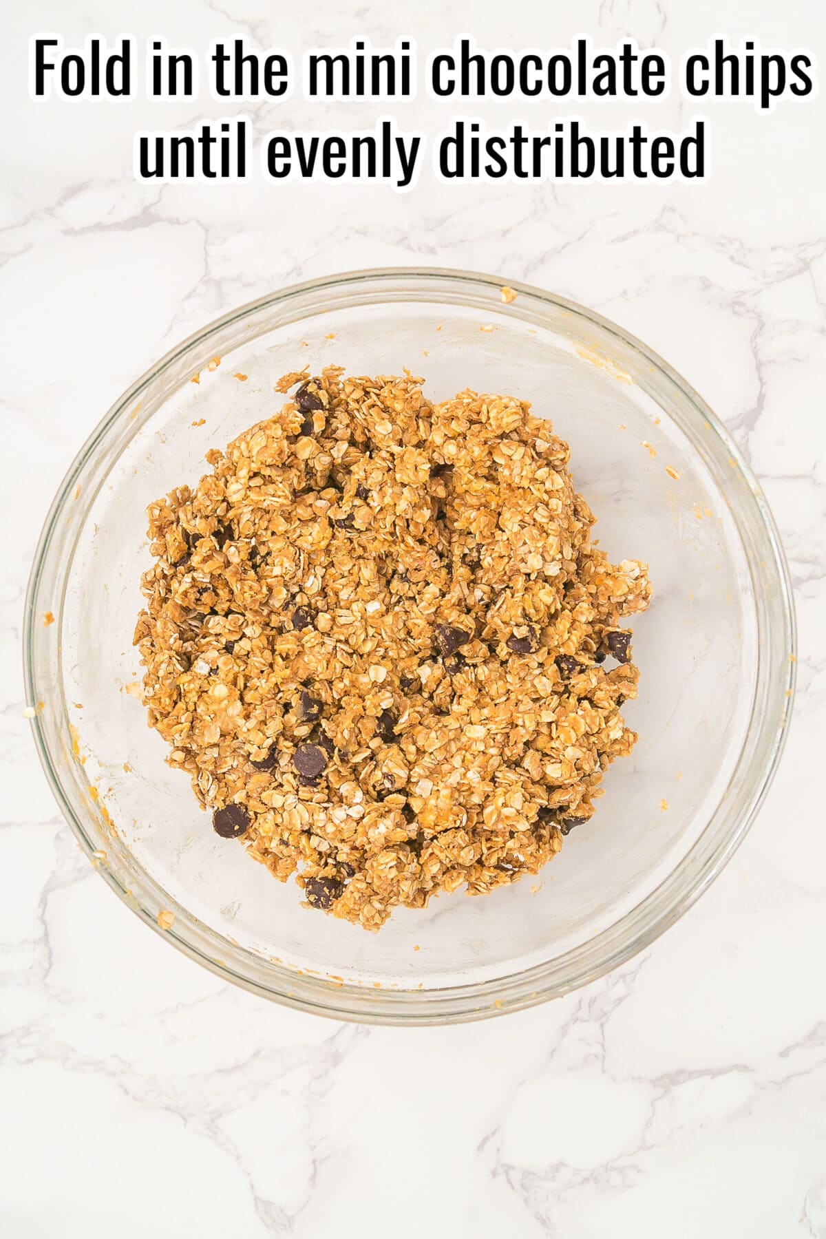 A glass bowl of oat and chocolate chip mixture on a marble surface with text instructing to fold in mini chocolate chips until evenly distributed.