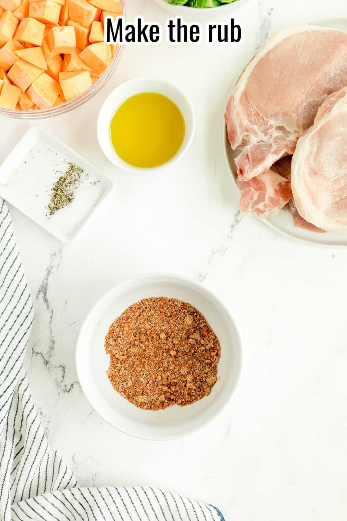 Overhead view of pork chops, a bowl of spice rub, cubed sweet potatoes, olive oil, salt, pepper, and a striped towel on a white surface. Text at top reads, “Make the rub.”.