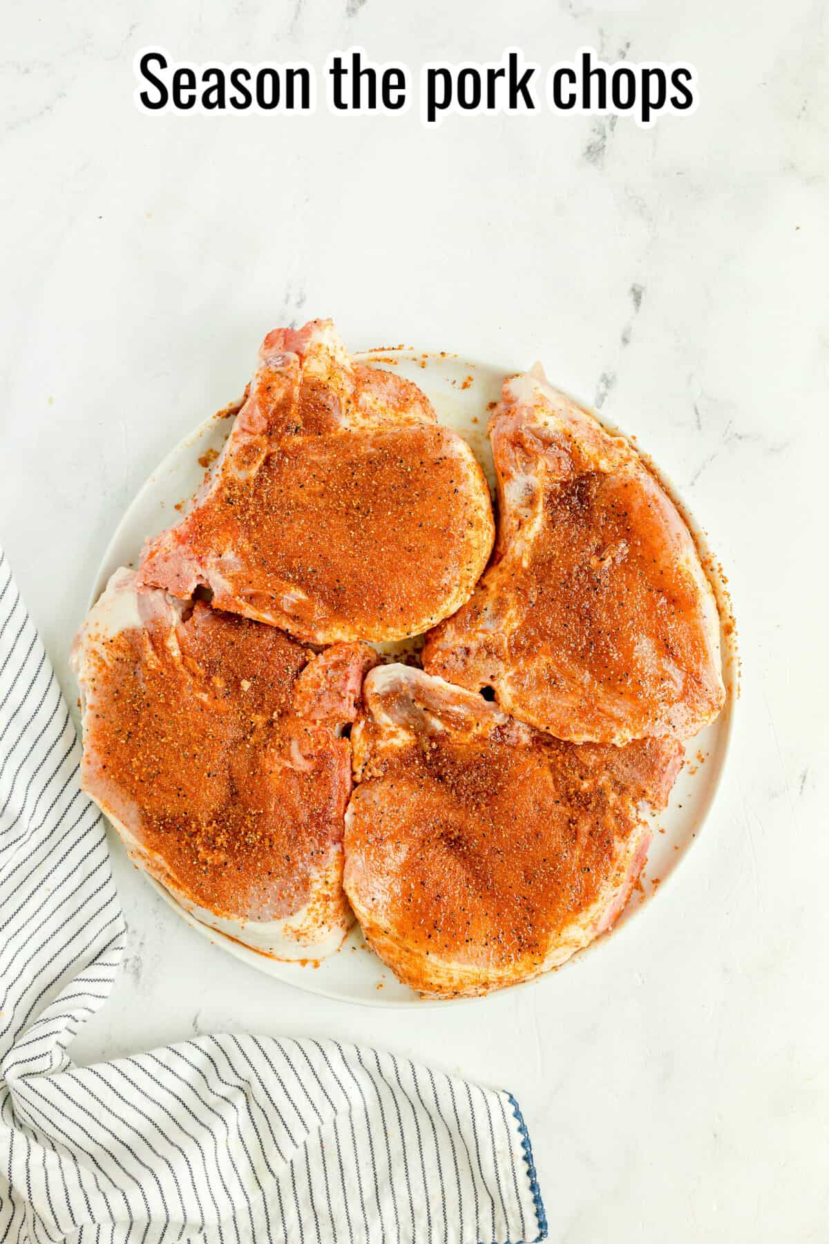 Four raw pork chops coated with seasoning are arranged on a white plate, set on a white surface beside a striped cloth. Text at the top reads, “Season the pork chops.”.
