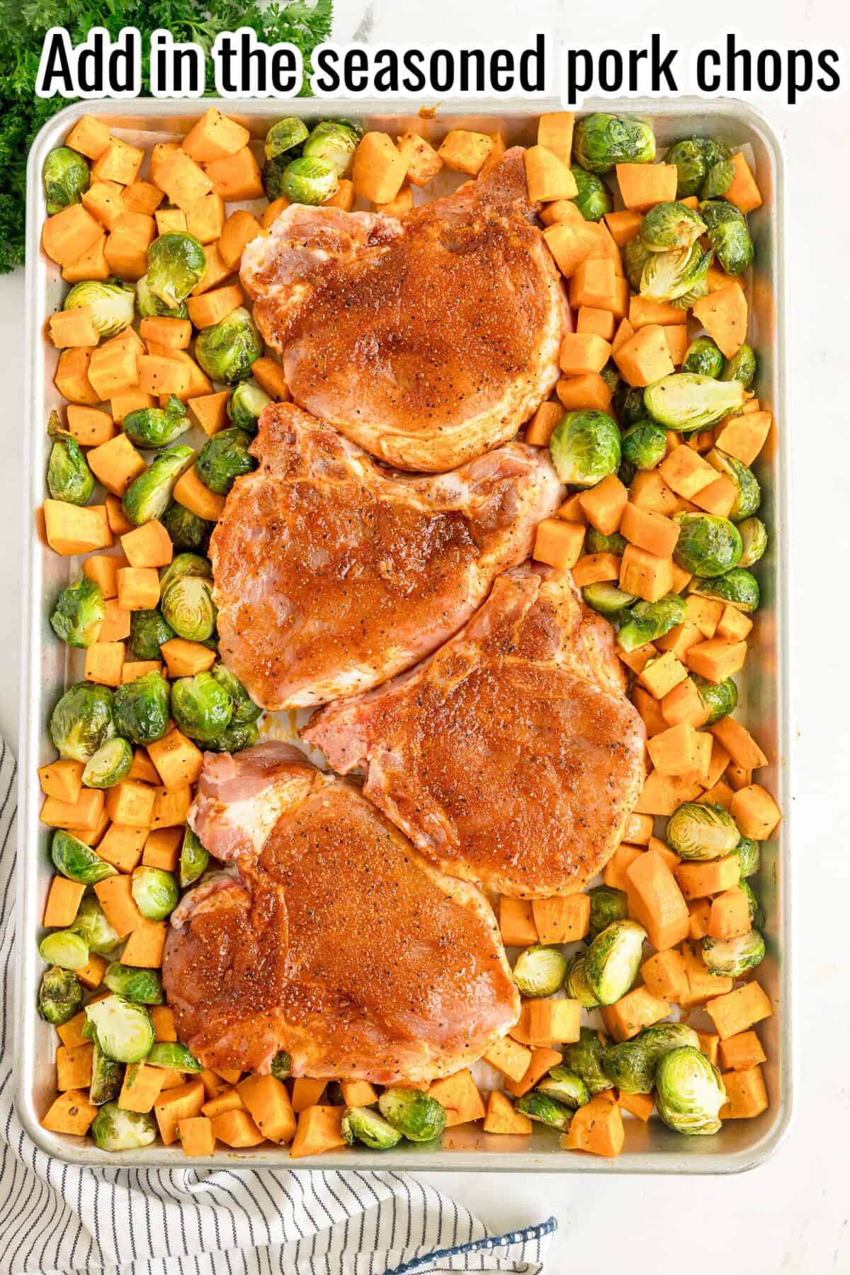Four seasoned pork chops on a baking sheet surrounded by chopped sweet potatoes and Brussels sprouts, ready for roasting.