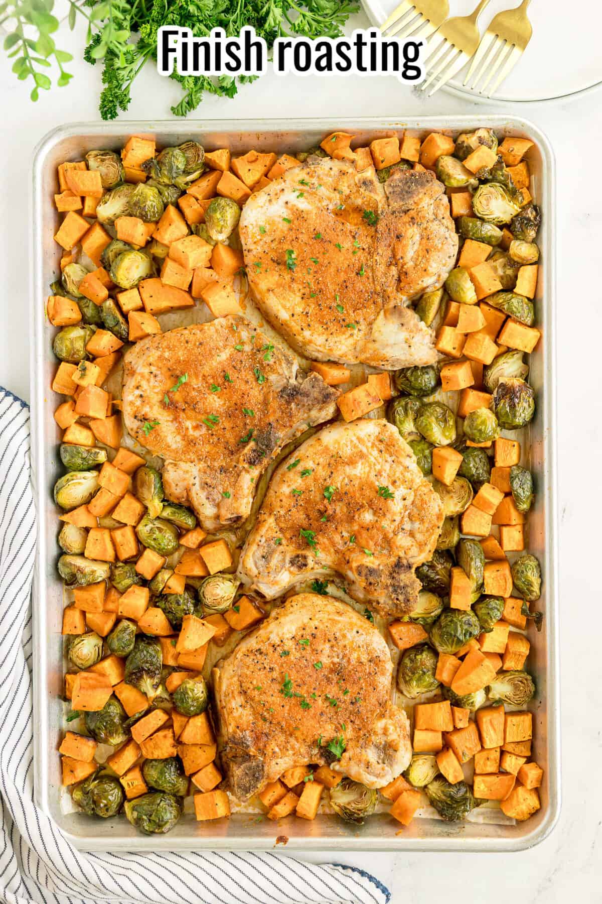 Sheet pan with four seasoned pork chops, diced sweet potatoes, and Brussels sprouts, all roasted. A napkin, greenery, and utensils are nearby. Text at the top reads “Finish roasting.”.