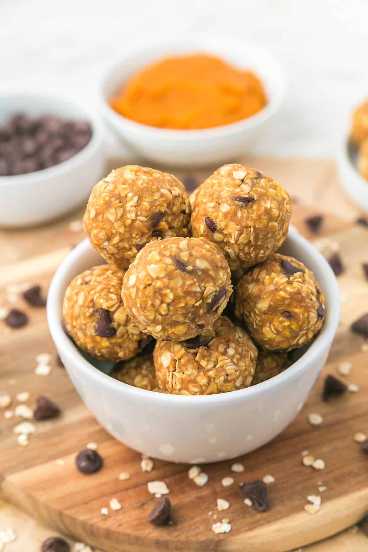 A white bowl filled with oat and chocolate chip energy bites sits on a wooden board, with scattered oats and chocolate chips around, and two ingredient bowls in the background.