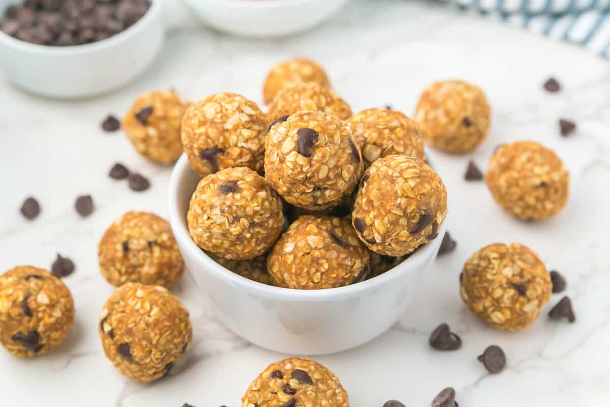 A white bowl filled with round pumpkin energy bites with chocolate chips, placed on a white marble surface with extra bites and chocolate chips scattered around.