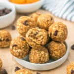 A small white plate holds a stack of oatmeal chocolate chip energy bites, with one bite taken to show the chewy texture. Other bites and chocolate chips are scattered around the plate.