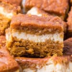 A square of pumpkin cheesecake bar with a layer of cream cheese filling sandwiched between two layers of pumpkin spice cake.