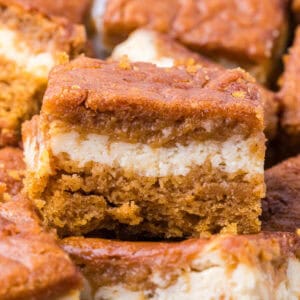 A square of pumpkin cheesecake bar with a layer of cream cheese filling sandwiched between two layers of pumpkin spice cake.