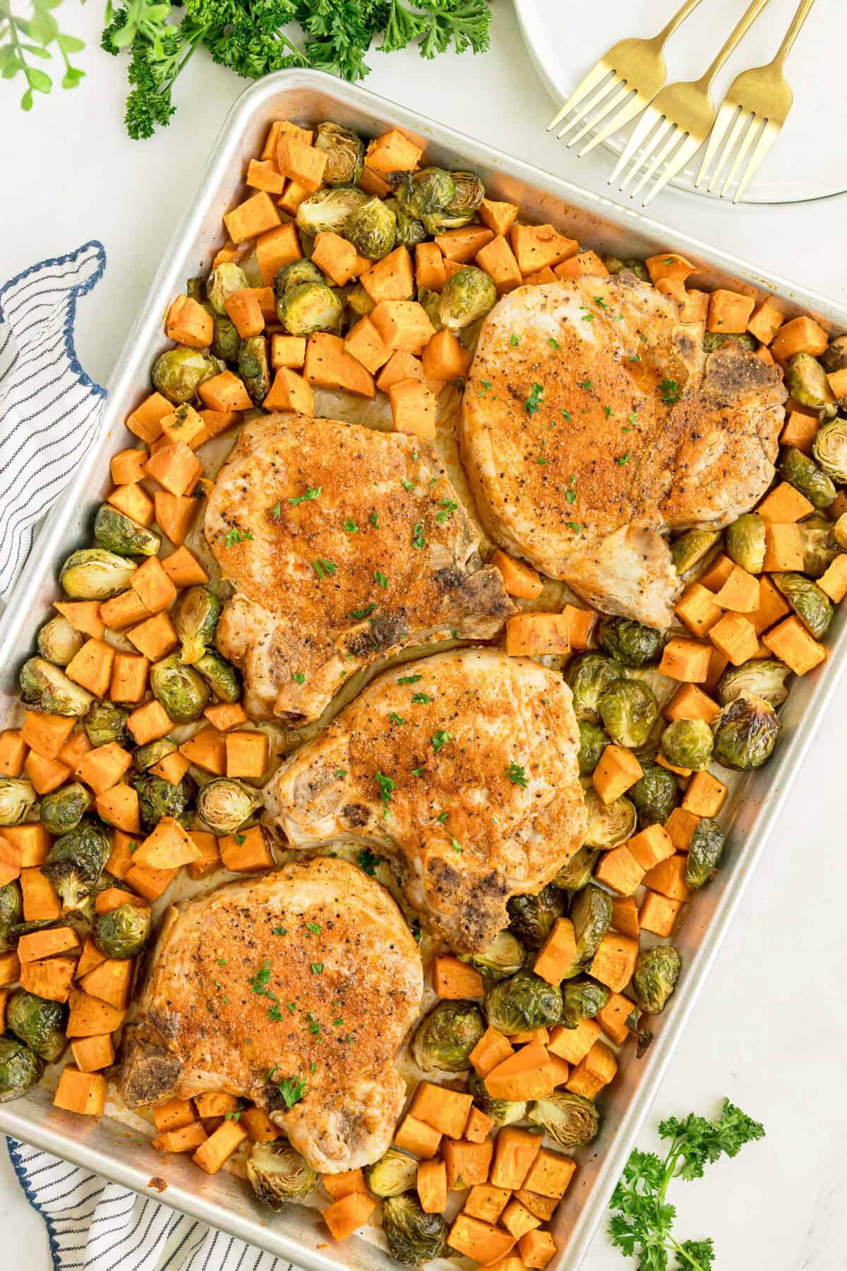 Sheet pan with four seasoned pork chops, diced sweet potatoes, and brussels sprouts, garnished with herbs. Plates, forks, and parsley are visible in the background.