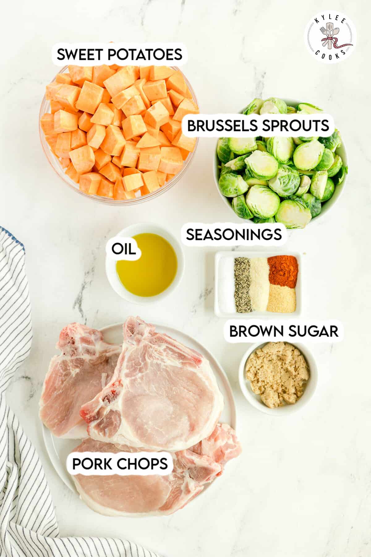 Ingredients for a meal are arranged on a white surface: sweet potatoes, Brussels sprouts, pork chops, oil, seasonings, and brown sugar, each in separate bowls or plates.