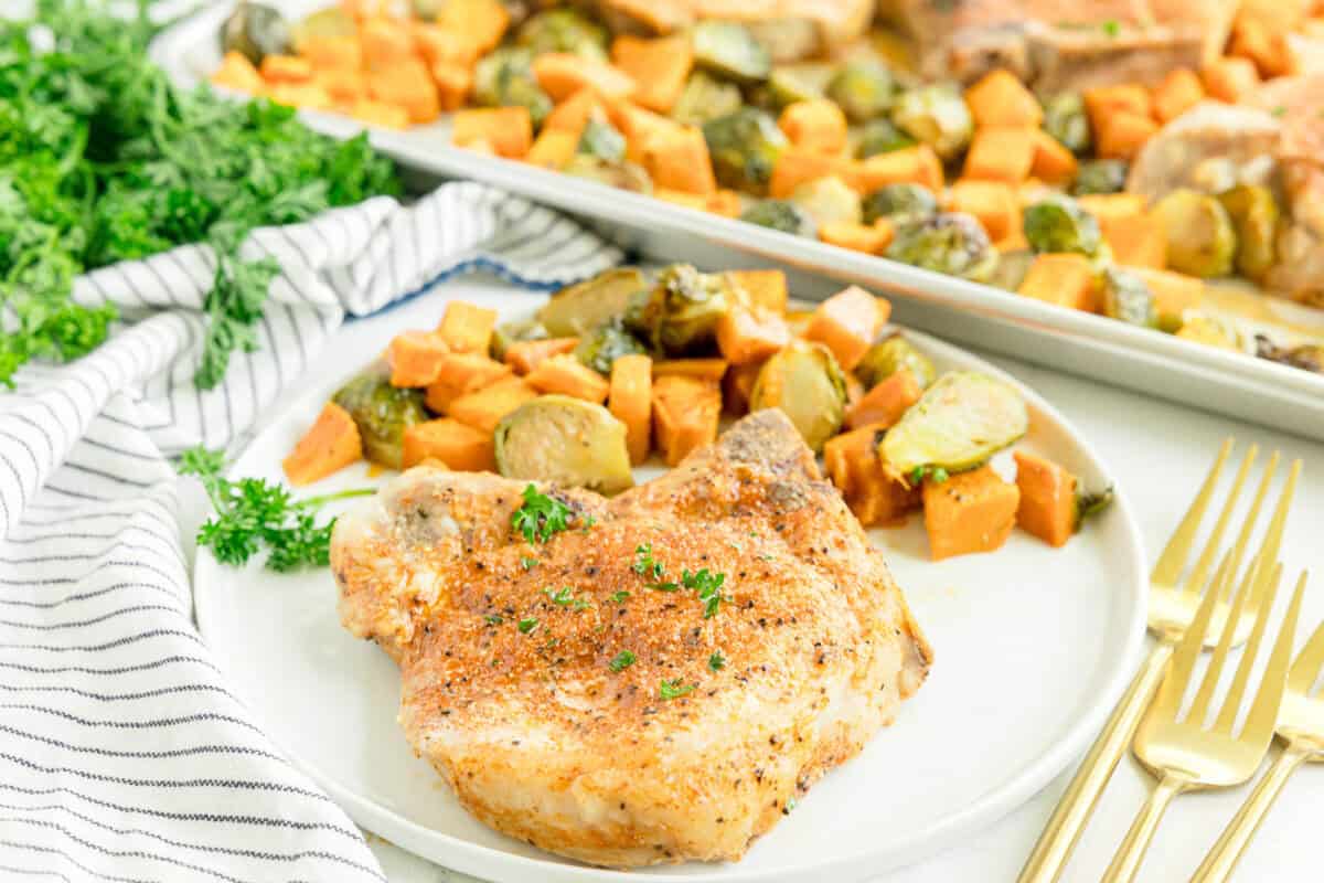 A seasoned pork chop with roasted sweet potatoes and Brussels sprouts on a white plate, with a fork, knife, and spoon beside it.