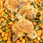 Three seasoned pork chops on a sheet pan, surrounded by roasted Brussels sprouts and diced sweet potatoes, garnished with chopped herbs.