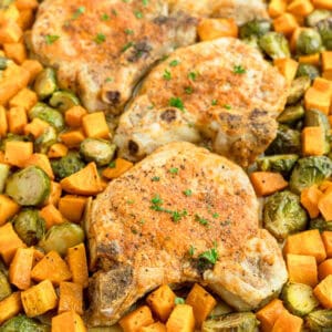 Three seasoned pork chops on a sheet pan, surrounded by roasted Brussels sprouts and diced sweet potatoes, garnished with chopped herbs.